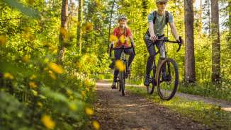 Twee fietsers met helmen rijden ontspannen op hun gravelfiets over een zonnig bospad door een groen bladerdak - actieve vakantie in de natuur van Liechtenstein