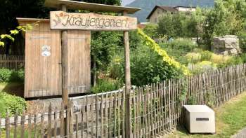 Houten bord met het opschrift "Kräutergarten" (kruidentuin) bij de ingang van de liefdevol aangelegde tuin in Mauren, omgeven door natuur en bergen.