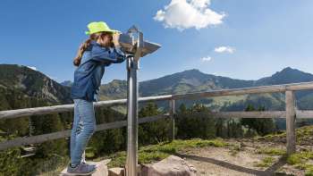 Meisje met groene hoed kijkt door een telescoop op het Forscherpad Malbun met een panoramisch uitzicht op de Liechtensteinse Alpen.