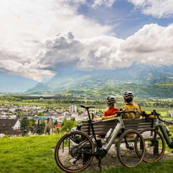 Twee fietsers met helmen op zitten op een bankje boven de Rijnvallei en genieten van het uitzicht op de vallei en de omliggende bergen - een gravel bike ervaring in Liechtenstein