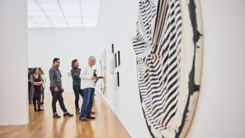 Groep bezoekers in de tentoonstellingsruimte van het kunstmuseum, op de voorgrond een groot, rond kunstwerk