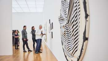 Groep bezoekers in de tentoonstellingsruimte van het kunstmuseum, op de voorgrond een groot, rond kunstwerk