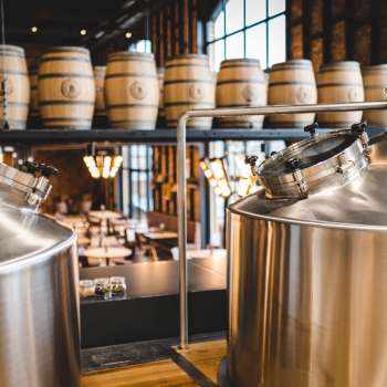 Zicht op een moderne brouwerij met glimmende roestvrijstalen tanks en gestapelde houten vaten op de achtergrond.