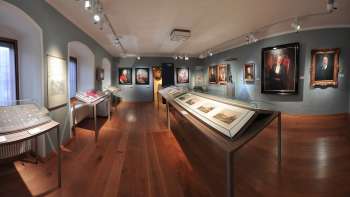 Panoramisch uitzicht in een tentoonstellingszaal van het Nationaal Museum van Liechtenstein met historische schilderijen en vitrines.