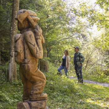 Twee wandelaars ontdekken een gebeeldhouwde figuur op het "pad van de Walser legendes" midden in een met licht overstroomd bos