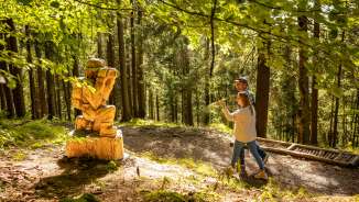 Twee bezoekers ontdekken een gesneden houten figuur langs de "pad van de Walser legendes" in Triesenberg, Liechtenstein - mystiek themawandelpad in het bos