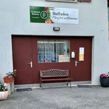 Ingang van de boerderijwinkel Aeulehof met rood hek, bankje en bloemdecoraties in Triesen, Liechtenstein.