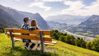 Echtpaar zittend op een bankje in Triesenberg en uitkijkend over de Rijnvallei in Liechtenstein met een indrukwekkend Alpenpanorama