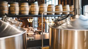 Zicht op een moderne brouwerij met glimmende roestvrijstalen tanks en gestapelde houten vaten op de achtergrond.