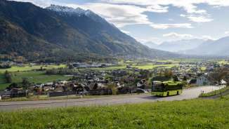Groene LIEmobil-bus rijdt over een kronkelige weg boven Liechtenstein met uitzicht op het Rijndal en de omringende Alpen