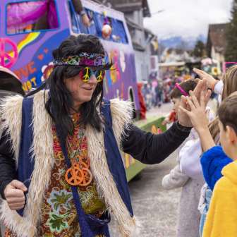 Hippie verkleed op het kindercarnaval in Mauren, high-fives met kinderen voor een kleurrijke praalwagen.