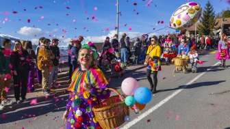 Clown verkleed met confetti en ballonnen tijdens de carnavalsoptocht, toeschouwers langs de kant van de weg.