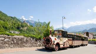 Citytrain rijdt door Vaduz, op de achtergrond het kasteel van Vaduz en de Alpen in de felle zon