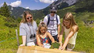 Familie verkent een interactief station op het themapad in Malbun, omgeven door alpenlandschap en zomerweiden in Liechtenstein.