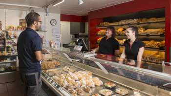 Verkoopruimte van een bakkerij met vriendelijk personeel, vers gebakken brood en gebak op de voorgrond