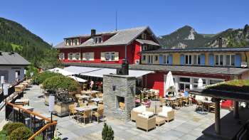 Buitenaanzicht van het restaurant in de zon, met rode gevel en bergachtig panorama.