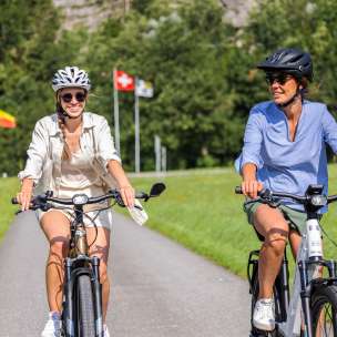 Twee vrouwen op e-bikes tijdens een zomerse tocht met de vlaggen van Liechtenstein, Zwitserland en Oostenrijk op de achtergrond