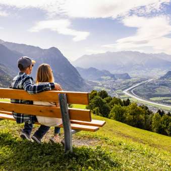 Echtpaar zittend op een bankje in Triesenberg en uitkijkend over de Rijnvallei in Liechtenstein met een indrukwekkend Alpenpanorama