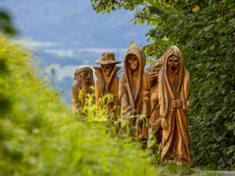 Detailopname van gebeeldhouwde houten figuren in mystieke uitbeelding op het Walser Legendepad in Triesenberg, omgeven door groene natuur