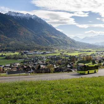 Groene LIEmobil-bus rijdt over een kronkelige weg boven Liechtenstein met uitzicht op het Rijndal en de omringende Alpen