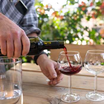 Een glas rode wijn wordt uit een fles in een wijnglas geschonken, met bloeiende planten op de achtergrond.