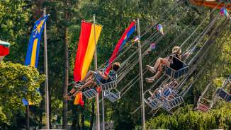 Kinderen en volwassenen rijden op een kettingcarrousel in de zon op de kermis in Balzers, omringd door kleurrijke vlaggen.