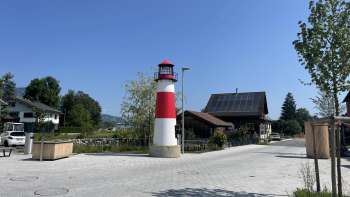 Kleine rood-witte vuurtoren van Moby op een plein in Ruggell bij zonnig weer, met traditionele gebouwen op de achtergrond.