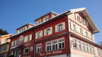 Buitenaanzicht van het Alpenhotel Vögeli Malbun met de Elchbar op de begane grond - traditioneel rood gebouw met witte letters tegen een blauwe lucht.