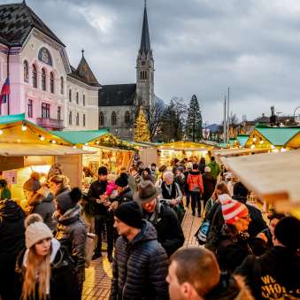 Sfeervolle avondopname van de kerstmarkt in Vaduz 2018 met feestelijk versierde marktkramen en mensenmassa's voor het regeringsgebouw en de kathedraal