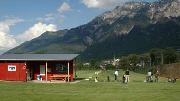 Golfers op de driving range in Liechtenstein met uitzicht op groene weiden, een rood clubhuis en het omringende berglandschap onder een blauwe hemel.