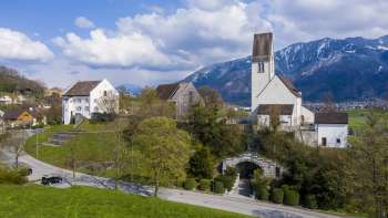 Panoramisch uitzicht op de "Kirchhügel" (kerkheuvel) van Bendern met zijn historische kerk, omgeven door goed onderhouden paden, bomen en traditionele architectuur tegen een alpiene bergachtergrond.