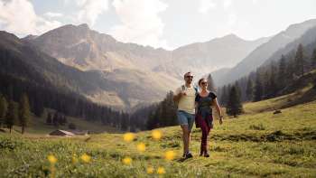 Twee mensen wandelen door een brede alpenweide met uitzicht op de omliggende bergen van de Valüna Alp bij zonnig weer