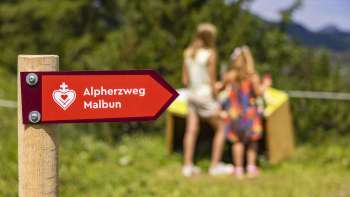 Wegwijzers van de Malbun Alpine Heart Trail met kinderen voor een van de stations op de achtergrond