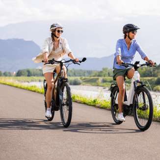 Twee vrouwen op e-bikes op het Rijnfietspad bij Liechtenstein, met rivier, groen landschap en bergen op de achtergrond