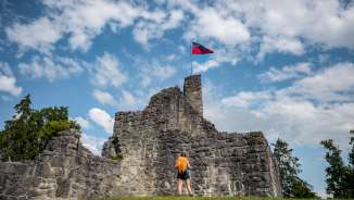 Wandelaar voor de historische ruïne van kasteel Schellenberg met de Liechtensteinse vlag onder een blauwe hemel