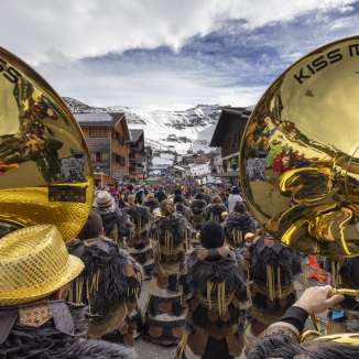Gezicht op een Guggenmusikgroep met gouden instrumenten tijdens de carnavalsoptocht in Malbun tegen een besneeuwde bergachtergrond.