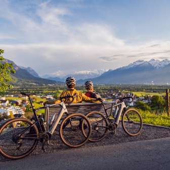 Twee fietsers met helmen op zitten op een bankje boven de Rijnvallei en genieten van de zonsondergang met uitzicht op de Alpen - hoogtepunt van een fietstocht in Liechtenstein