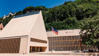 Modern parlementsgebouw in Vaduz met een puntgevel, de Liechtensteinse vlag ervoor en omgeven door groene heuvels.
