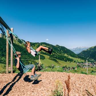 Twee kinderen schommelen op een speelplaats in de bergen met uitzicht op het zomerpanorama van Malbun in Liechtenstein