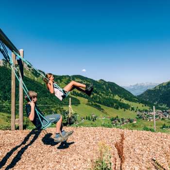 Twee kinderen schommelen op een speelplaats in de bergen met uitzicht op het zomerpanorama van Malbun in Liechtenstein