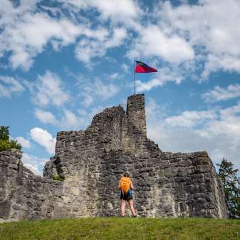 Wandelaar voor de historische ruïne van kasteel Schellenberg met de Liechtensteinse vlag onder een blauwe hemel