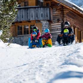 Twee volwassenen en drie kinderen zitten op sleeën en beginnen samen aan een afdaling voor een besneeuwde berghut in Malbun