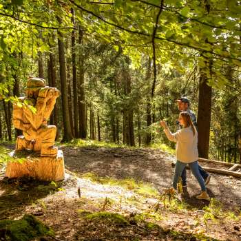 Vrouw wijst naar een gedetailleerd houten beeld in het bos op "pad van de Walser legendes".