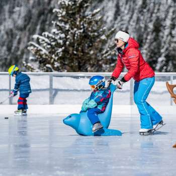 Familie op de ijsbaan bij Schlucher-Treff Malbun, ondersteund door een schaatshulp in de vorm van een blauw dier.