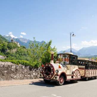 Citytrain rijdt door Vaduz, op de achtergrond het kasteel van Vaduz en de Alpen in de felle zon