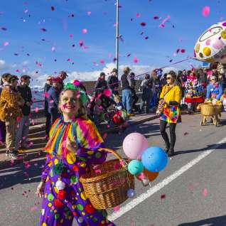 Clown verkleed met confetti en ballonnen tijdens de carnavalsoptocht, toeschouwers langs de kant van de weg.