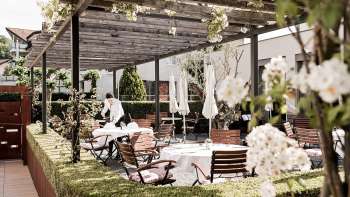 Een elegant terras met witte tafels gedekt met servetten en glazen onder een houten paviljoen, omringd door weelderig groen.
