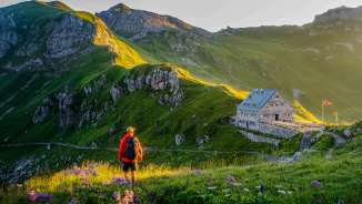 Wandelaar kijkt naar de Pfälzerhütte in Liechtenstein, omgeven door een groen berglandschap en Alpenpanorama in het avondlicht