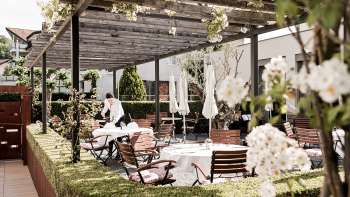 Een elegant terras met witte tafels gedekt met servetten en glazen onder een houten paviljoen, omringd door weelderig groen. Een elegant terras met witte tafels gedekt met servetten en glazen onder een houten paviljoen, omringd door weelderig groen.