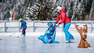 Moeder duwt kind op een schaatsende pinguïn over de ijsbaan in Malbun, op de achtergrond een kind met een ijshockeystick